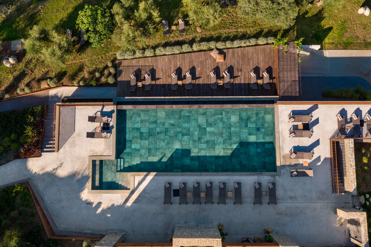 A drone view of a large turquoise infinity pool and its surrounding green trees and gardens and handful of sun loungers