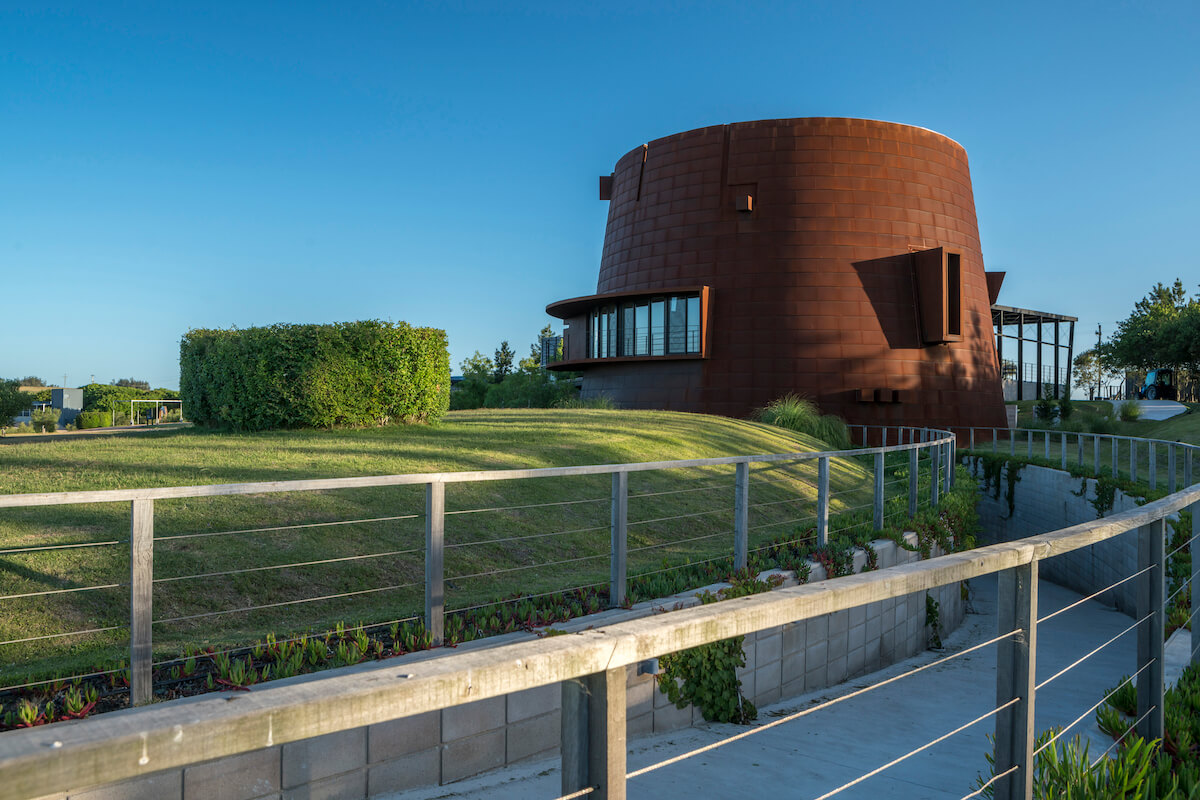 A sculptural wine cellar shaped like a cone against the bright blue sky and surrounding green grassland.